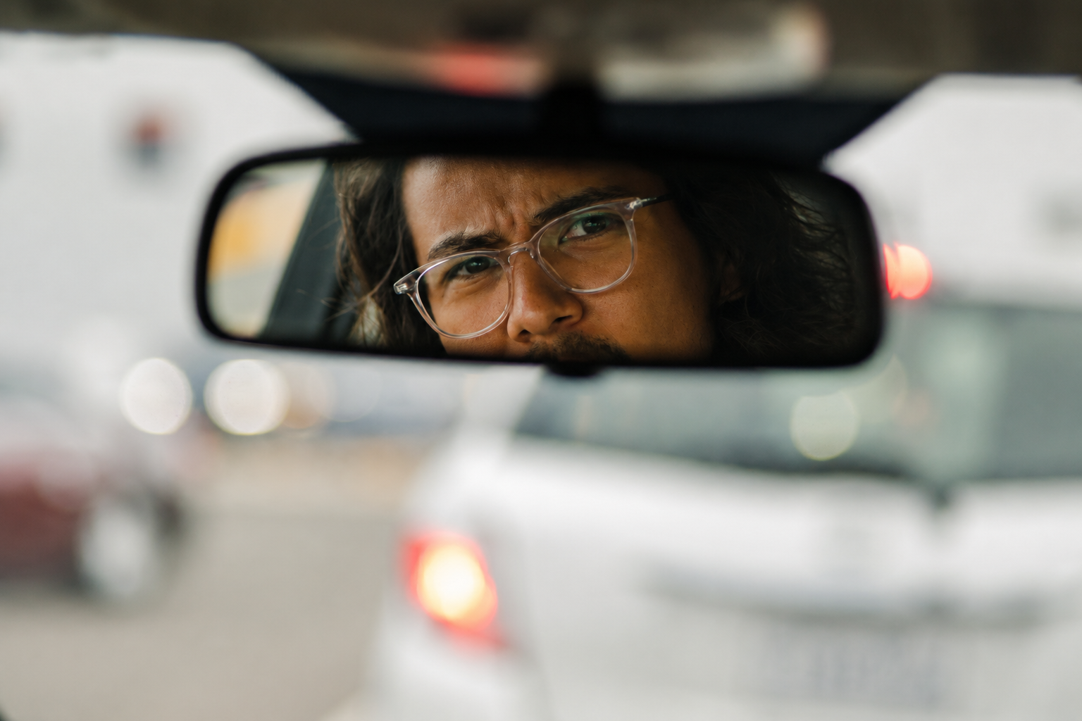 A person with glasses is seen in a car's rearview mirror, looking intently forward. Blurred traffic and a white car are visible in the background.