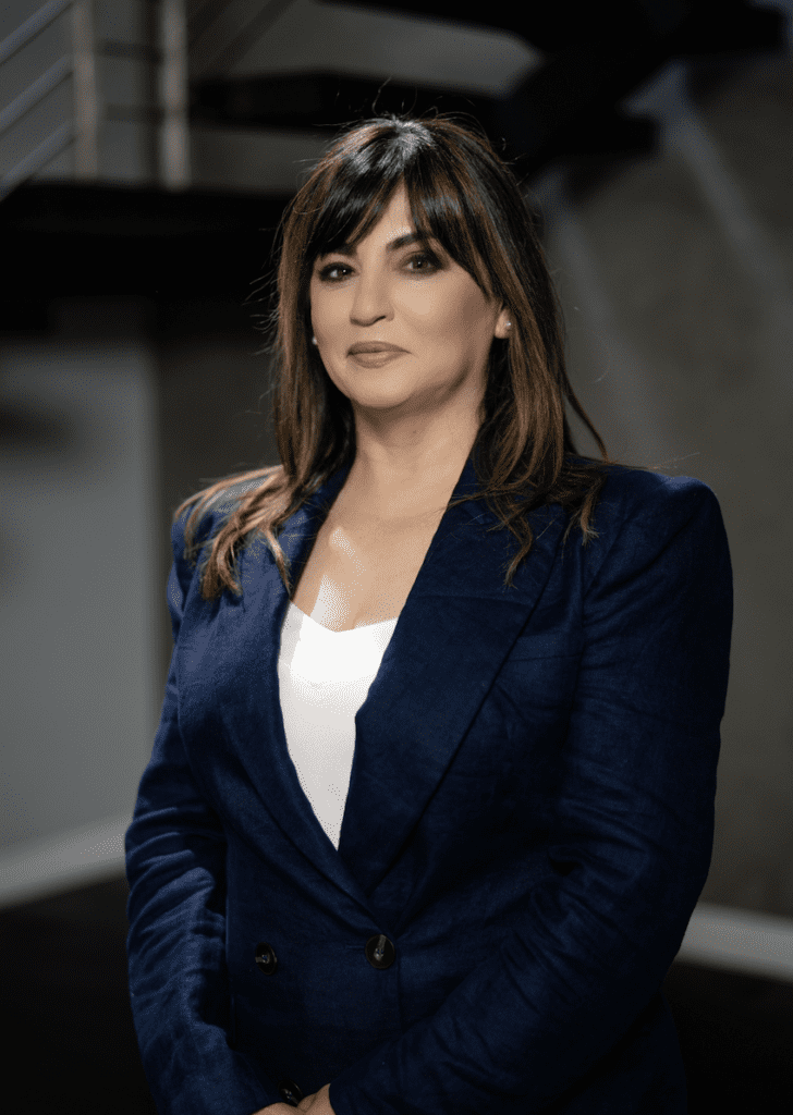 A woman with straight brown hair wearing a navy blazer and white top stands indoors, facing the camera with a neutral expression.