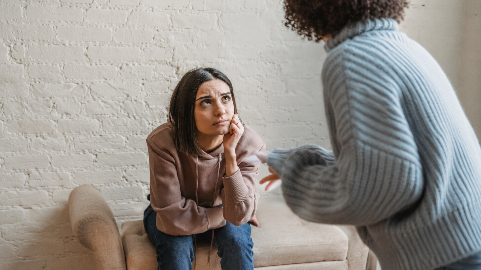 A woman sits on a bench, appearing concerned while listening to another person who is standing and speaking to her.