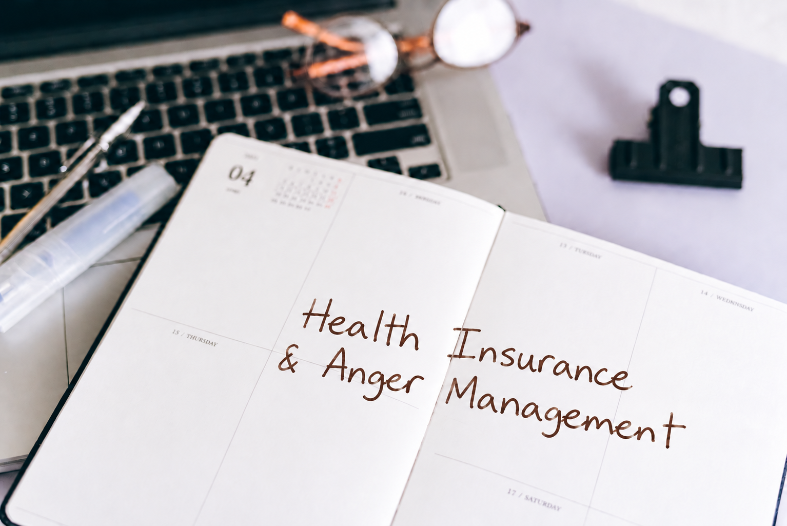 An open planner on a desk displays the words “Health Insurance & Anger Management” next to a laptop, glasses, and office supplies.